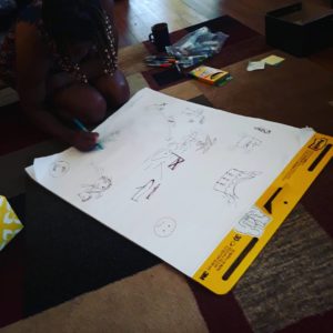 a Black woman sits on the floor drawing on a large piece of paper with multiple markers