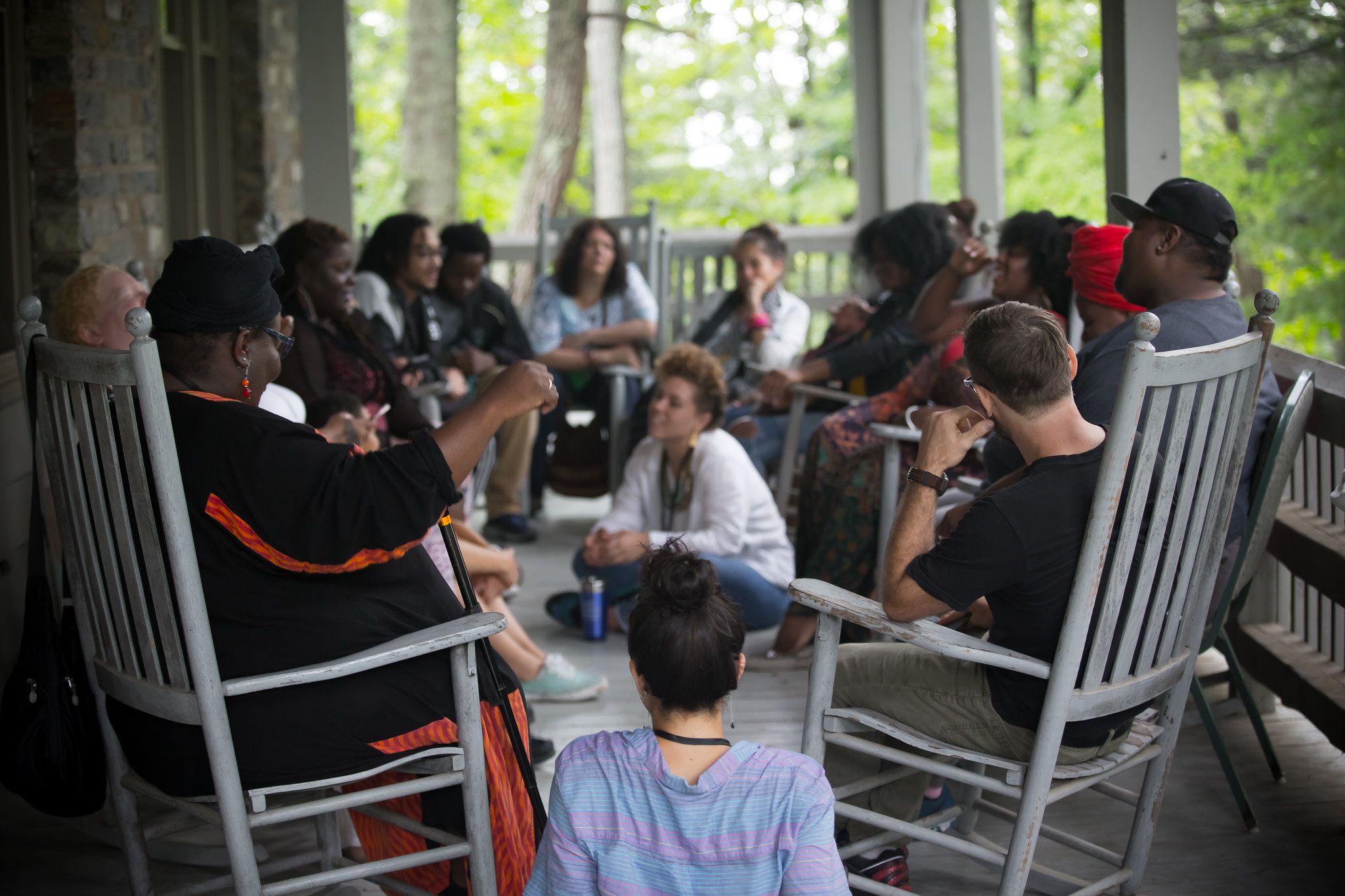 a group of many different kinds of people sit in a circle of rocking chairs on a porch in summer time, some are on the floor