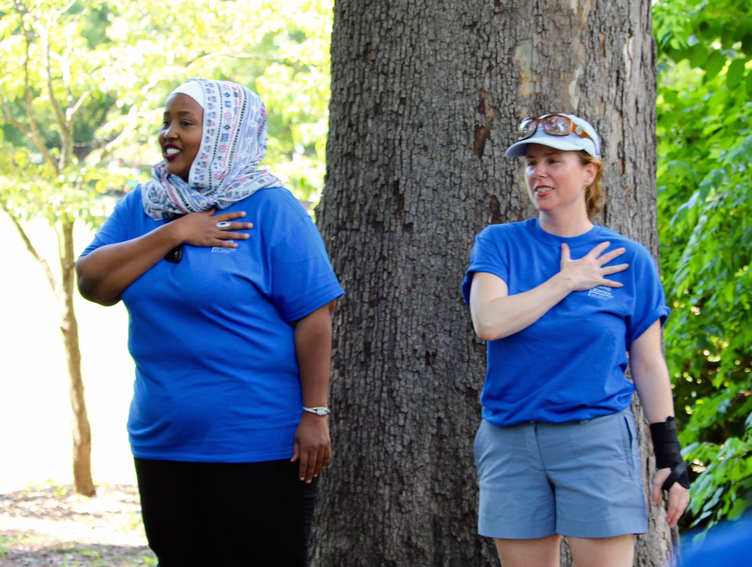 an African woman with a headscarf and a white woman wearing a ball cap are both crossing their hearts, each are wearing blue t-shirts