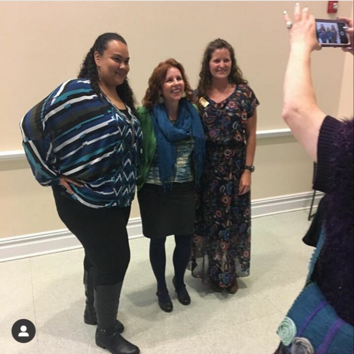 three women pose together for a woman taking a photo on a cellphone