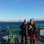 Two white women stand on the deck of a ferry with a bright blue sky and the the Seattle skyline far behind them. They are wearing heavy winter coats.