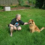 An older woman crouches down in a yard to pet a fluffy red dog with soulful eyes.